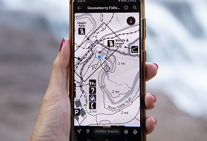 A hand holding a cellphone with a geoPDF showing a state park hiking trail, with the trail beckoning in the background